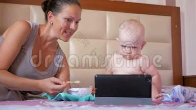 年轻的妈妈用一勺粥喂可爱的宝宝.. 宝宝坐在床上盯着平板电脑上的卡通片